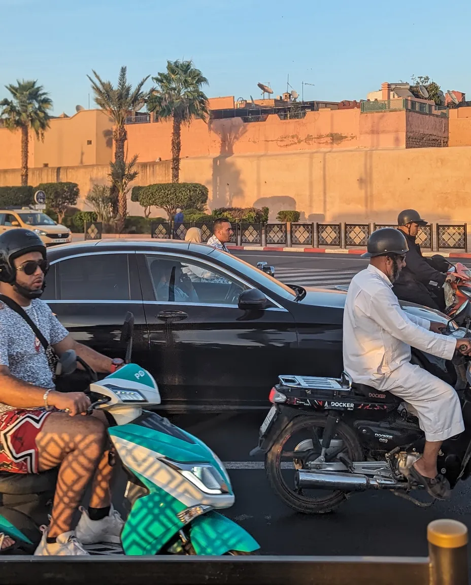 Evening traffic beside the medina walls in Marrakech — scooters, cars and pedestrians at a busy intersection
